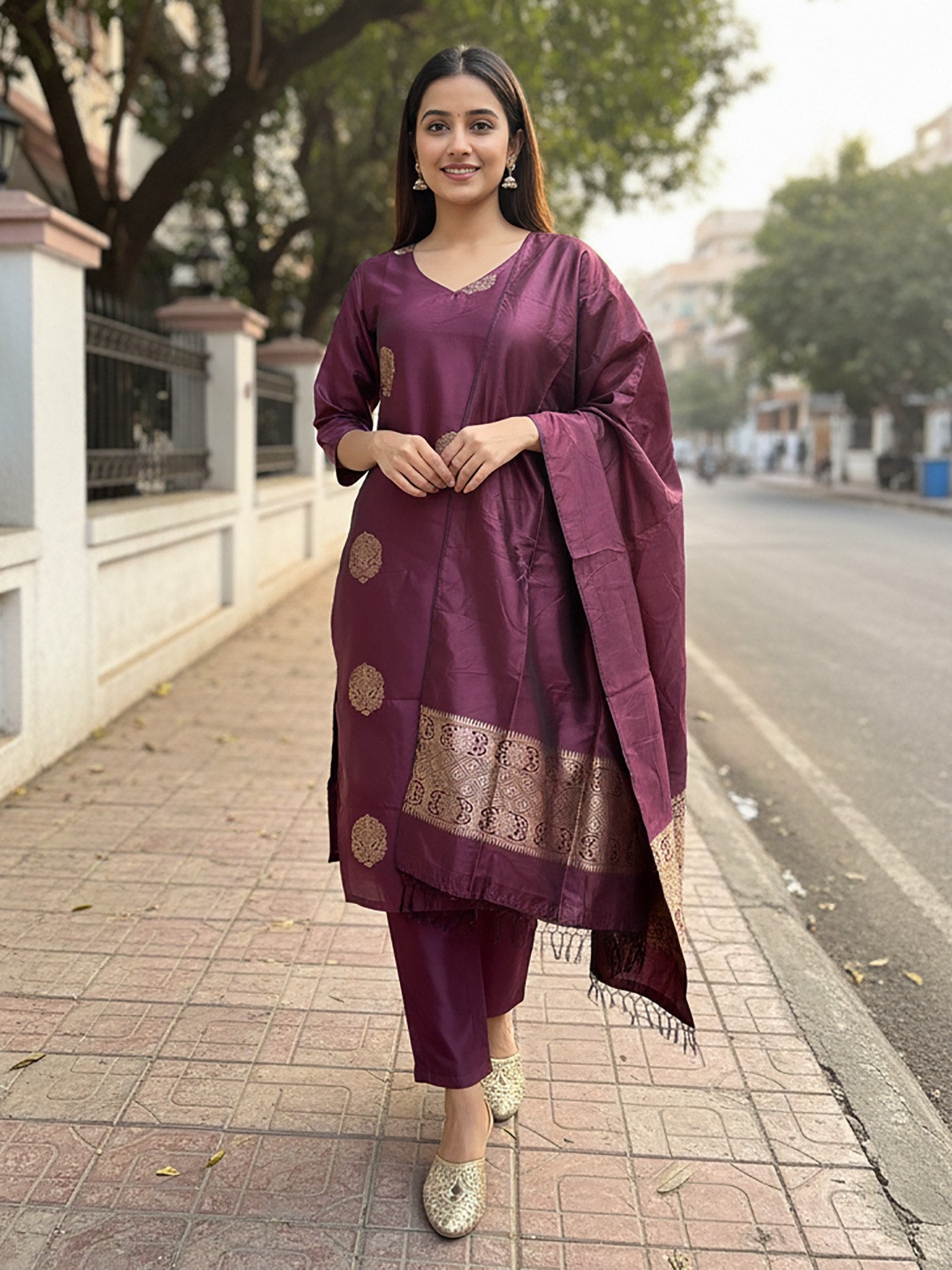 Woman wearing an aubergine silk blend kurta set with subtle gold woven motifs, straight pants, and a matching dupatta with a rich gold zari woven border.