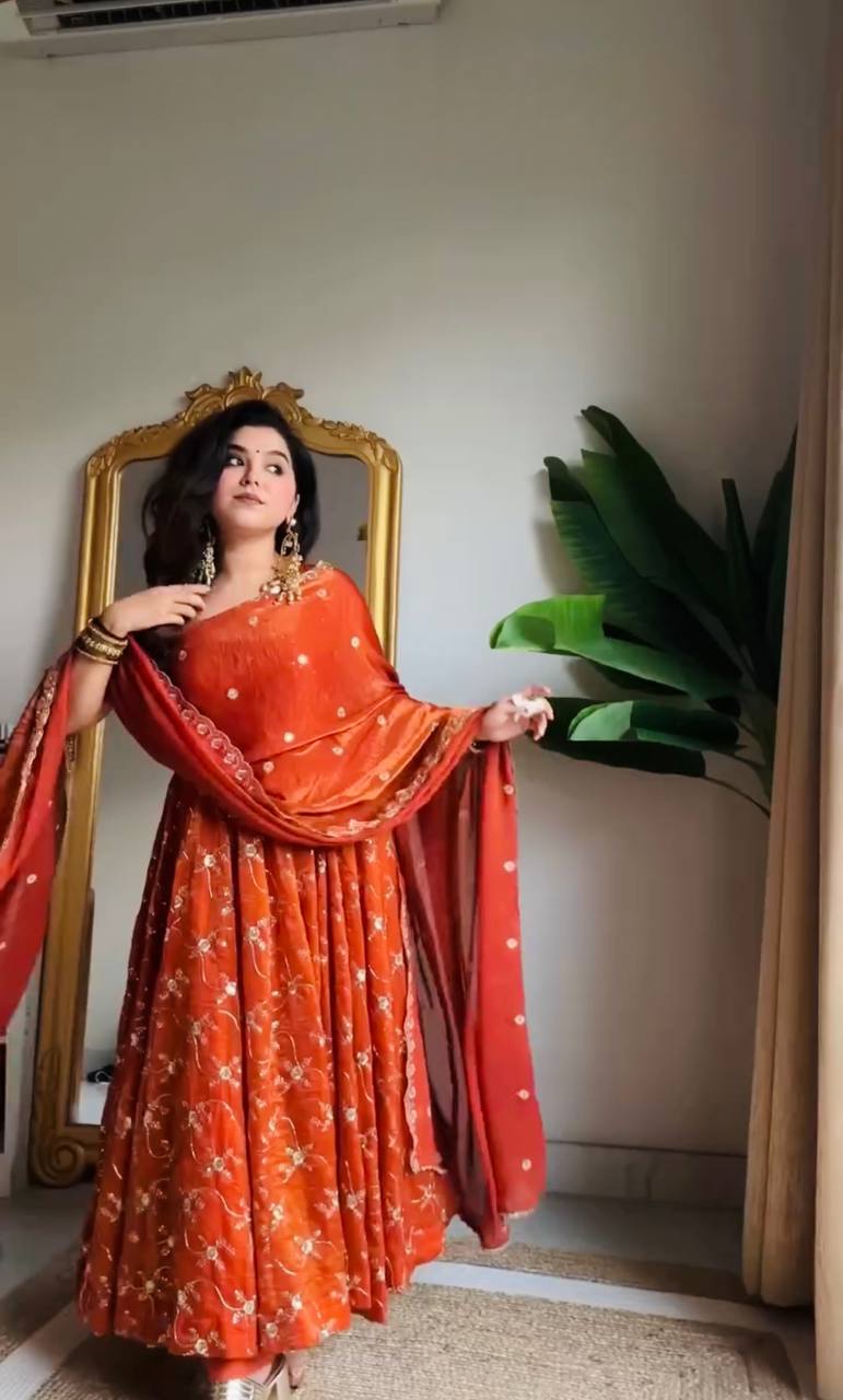 Woman wearing a fiery orange georgette anarkali gown with all-over gold floral print or embroidery, strappy sleeves, a generous flare, and a matching dupatta, styled with gold jewelry.