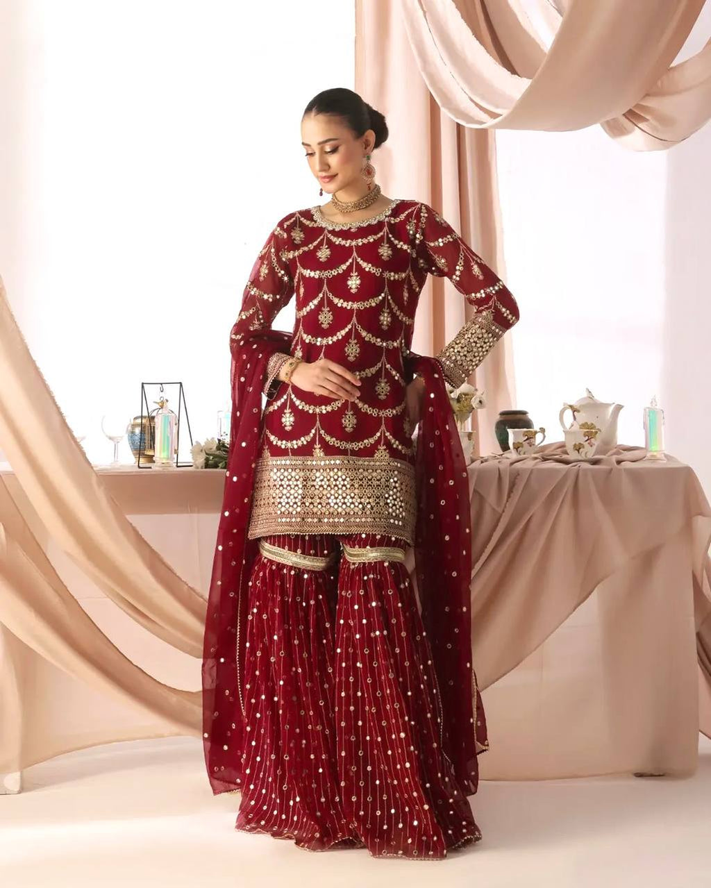 Woman wearing a deep maroon red georgette kurta sharara suit with heavy gold sequin and zari embroidery on the kurta, full sleeves, and tiered sharara, with a matching sheer dupatta.