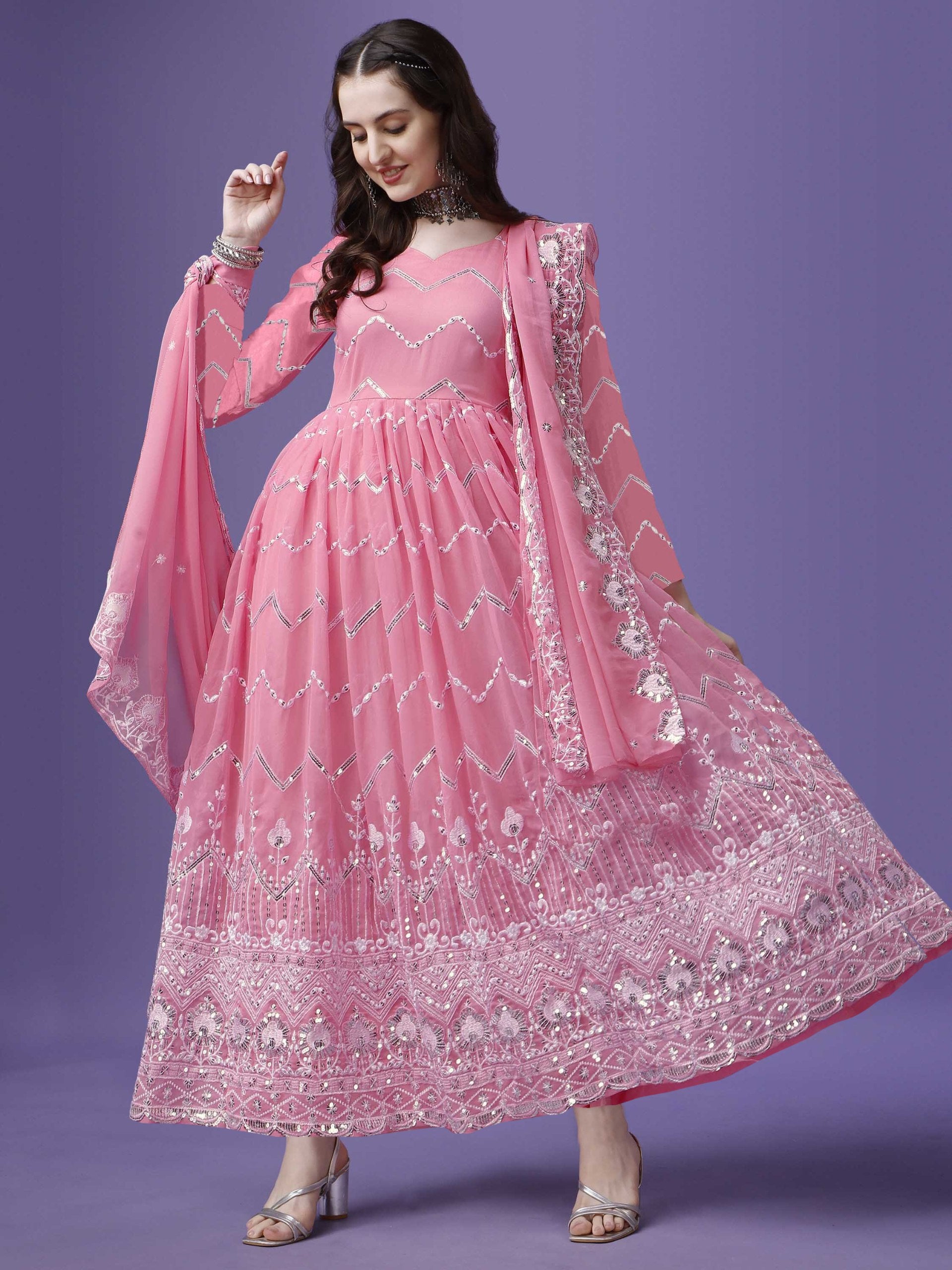 Woman wearing a flowing pink georgette anarkali gown with intricate silver zari and sequin embroidery on the bodice and flared hem, paired with a matching dupatta.
