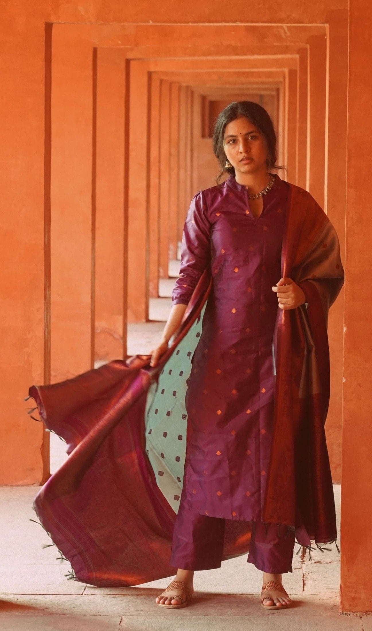 Woman wearing a plum silk blend kurta set with subtle motifs, straight pants, and a dual-tone woven dupatta with tassels.