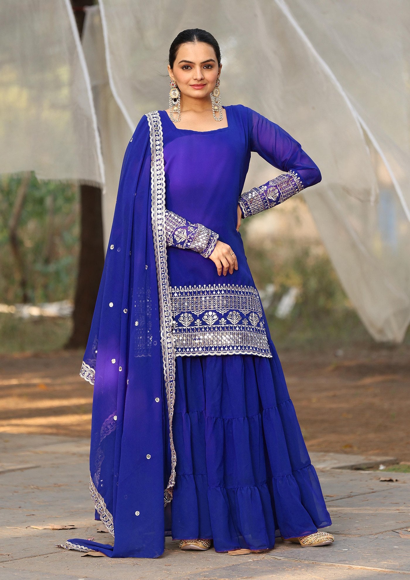 Woman wearing a royal blue georgette sharara suit with intricate silver zari embroidery on the full sleeves and kurta hem, paired with a voluminous tiered sharara and matching dupatta.