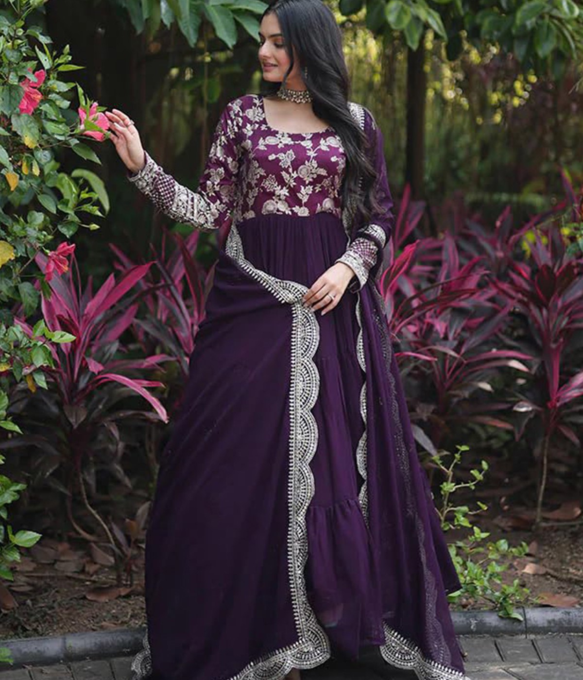 Woman wearing a royal purple georgette tiered anarkali gown with heavy gold zari embroidery on the bodice and sleeves, paired with a matching scalloped dupatta.