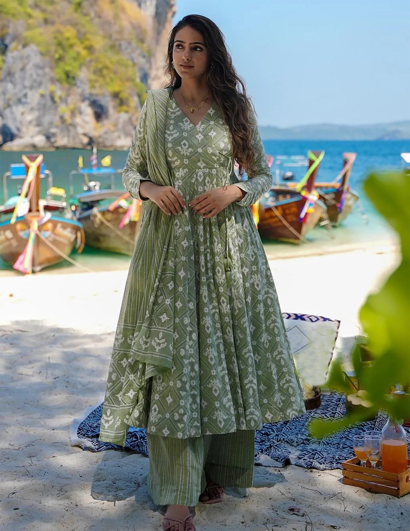 Woman wearing a sage green cotton printed anarkali kurta with full sleeves, paired with striped palazzo pants and a coordinating printed dupatta, on a beach setting.