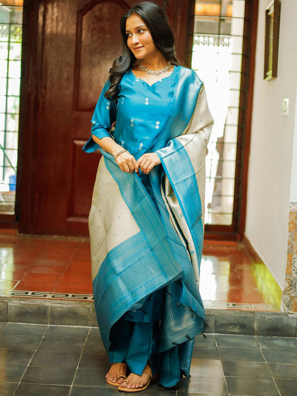 Woman wearing a teal blue silk kurta pant set with a heavy cream and gold Banarasi zari woven dupatta featuring a contrast blue border, styled with traditional gold jewelry.