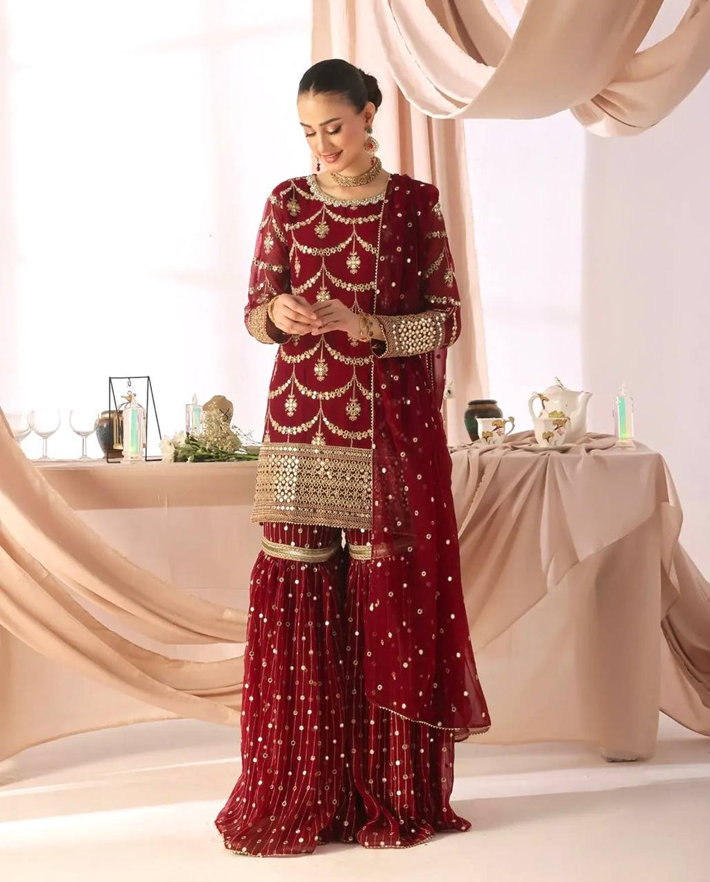 Model in a wedding guest red sharara suit with full sleeves, intricate gold sequin embroidery on the kurta, and a tiered sharara with scattered sequins.